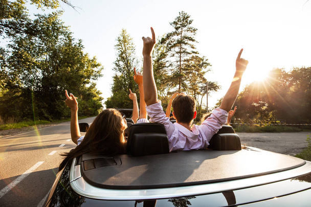 Company of young people riding in a cabriolet on the road and holding their hands up on a warm sunny day. Back view. - Foto, immagini