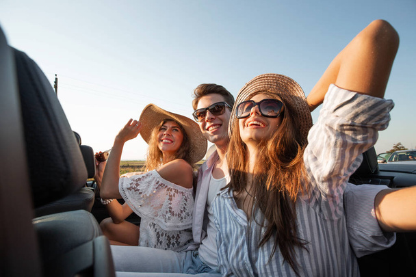 Charming young dark-haired women in hats are sitting with young  man in a black cabriolet and smiling on a summer day. - Foto, imagen