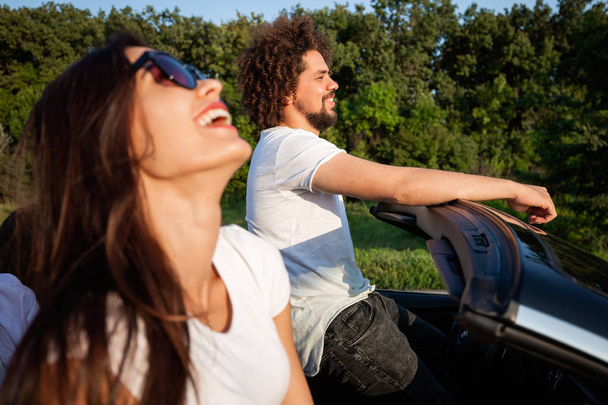 Gorgeous young dark-haired woman in sunglasses with curly young man are sitting and smiling in a black cabriolet on a sunny day. - Photo, Image