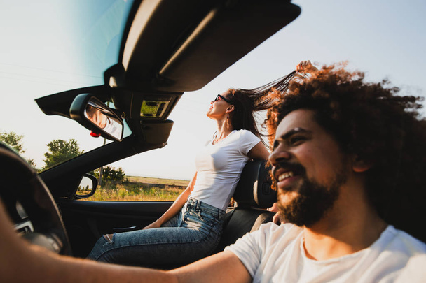 Göndör sötét hajú fiatalember ült a volánnál, egy fekete cabriolet és gyönyörű nő ül mellette egy napsütéses napon. - Fotó, kép