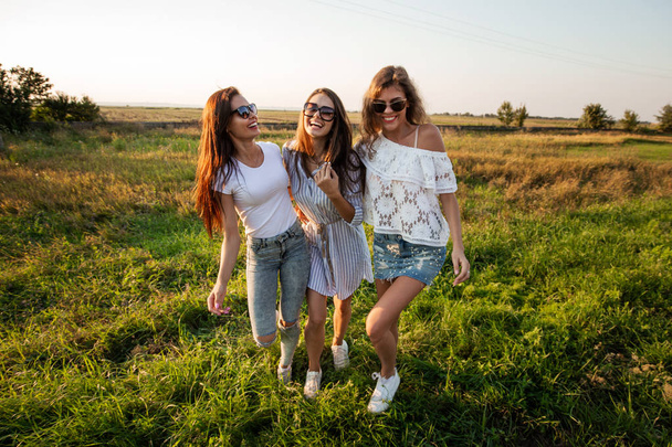 Three gorgeous young women in sunglasses dressed in the beautiful clothes stand in the field and smiling on a sunny day. - Zdjęcie, obraz