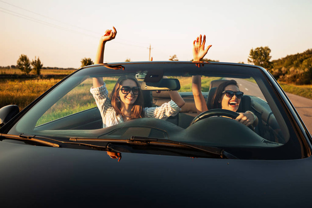 Two charming dark-haired young women in sunglasses are sitting in a black cabriolet on a sunny day. - Fotó, kép
