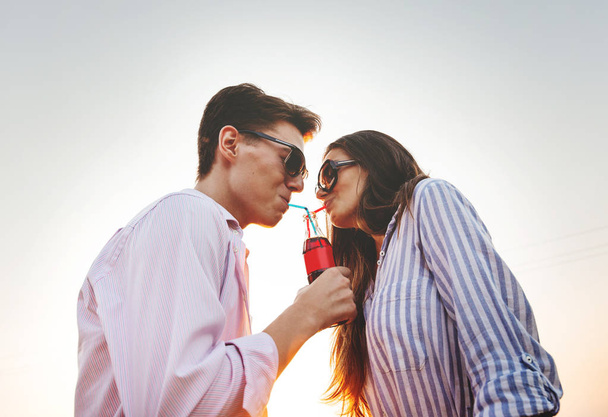 Beautiful dark-haired girl and a young man in sunglasses are drinking from one bottle a drink through straws outdoor on a sunny day. - Foto, Imagen