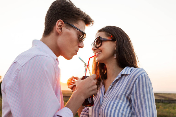 Beautiful dark-haired girl and a young man  in sunglasses are drinking from one bottle a drink through straws outdoor on a sunny day. - Φωτογραφία, εικόνα