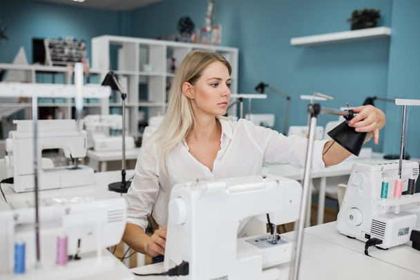 A smart-looking pretty blond woman wearing white shirt is sewing with the electric sewing-machine. Fashion, tailors workshop. - Photo, Image
