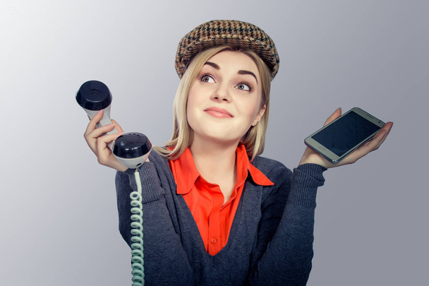 Young blond woman choosing between retro phone and modern smartphone on gray background - 写真・画像