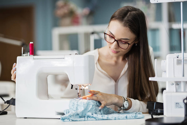 A smart-looking pretty brunet woman wearing white shirt is sewing with the electric sewing-machine. Fashion, tailors workshop. - Zdjęcie, obraz