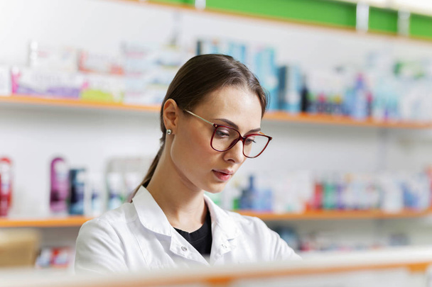 A good looking slim girl with dark hair and glasses,wearing a white coat, gently places goods on the shelf in the pharmacy. - Foto, immagini