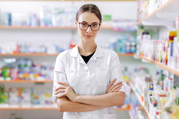 A young slim girl with long dark hair and glasses,wearing a white coat, confidently stands by shelves in a up-to-date pharmacy. - Photo, image