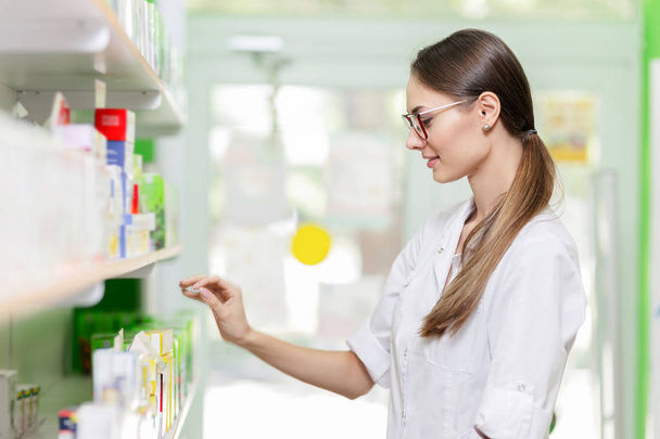 A good looking slim girl with dark hair and glasses,wearing a lab coat,takes something from the shelf in a new pharmacy. - Foto, immagini