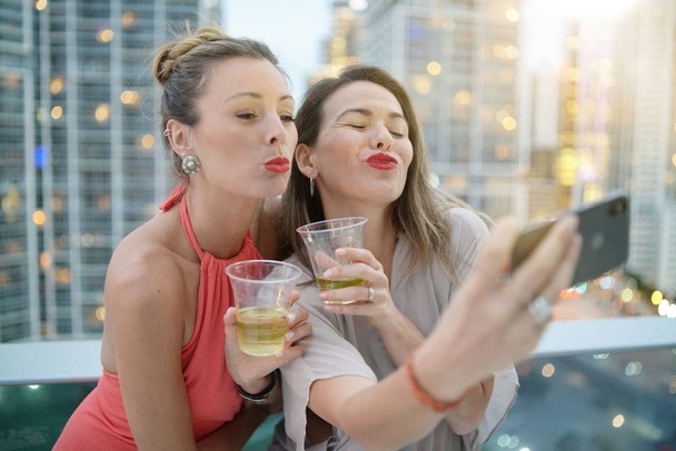Dos hermosos amigos tomando selfie en el bar de la azotea con luces de la ciudad telón de fondo
 - Foto, imagen