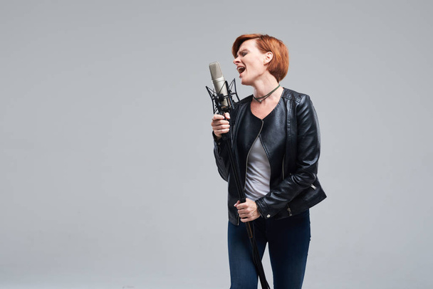 Portrait of rock singer wearing leather jacket and keeping static mic, sings a song loudly on grey background. Concept of rock music and rave - Foto, Imagem