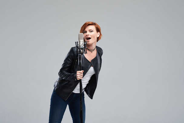 Portrait of rock singer wearing leather jacket and keeping static mic, sings a song loudly on grey background. Concept of rock music and rave - Photo, Image