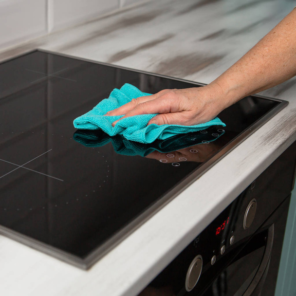 A woman's hand with a blue microfiber cloth rubs a glass ceramic stove in the kitchen. - Photo, image