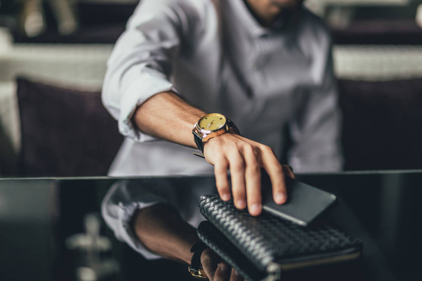 A businessmans hand with watch holding phone on a purse on a table in a cafe - Photo, Image