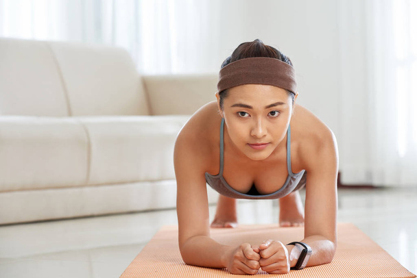 Jovem mulher asiática atraente em pé na posição de prancha e fortalecendo os músculos do abdômen enquanto exercita em casa
 - Foto, Imagem