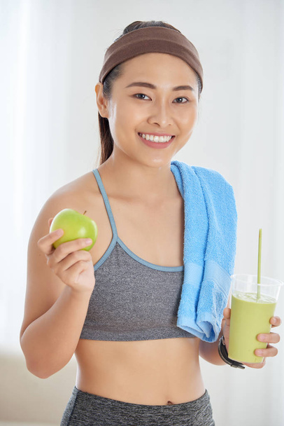 Portrait of young beautiful athletic Asian woman in active wear smiling at camera joyfully with smoothie and organic apple in her hands - Photo, Image