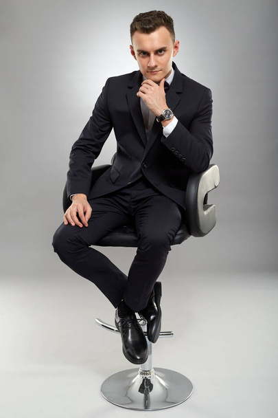 Young businessman sitting on chair, gray background - Φωτογραφία, εικόνα