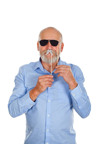Cool elderly man having fun with photo booth accessories. Holding black sunglasses and mustache on isolated background - Foto, Imagem
