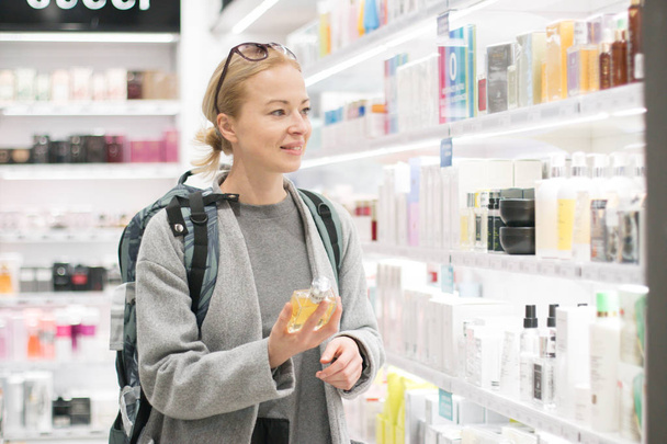 Bionda giovane viaggiatore donna che indossa zaino da viaggio scegliendo profumo in aeroporto duty free store
. - Foto, immagini