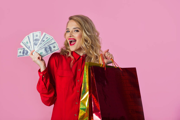 beautiful young girl in a red shirt with dollars and packages in their hands on a pink background - Foto, imagen