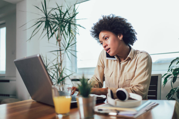 Lachende jonge Afrikaanse vrouwelijke ondernemer zit aan een bureau in haar kantoor aan huis werken online met een laptop - Foto, afbeelding