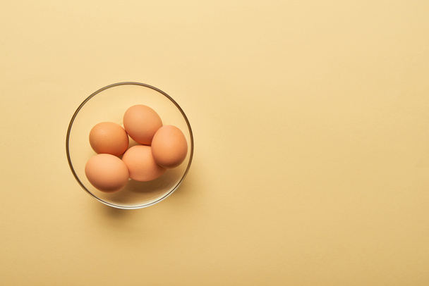 top view of fresh eggs in glass bowl on yellow background - Foto, imagen