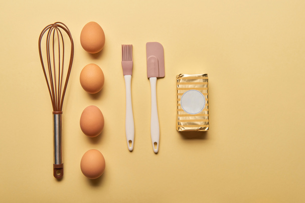 επίπεδη lay με χτυπητήρι, αυγά, σπάτουλα, βούρτσα αρτοποιίας και βούτυρο σε κίτρινο φόντο - Φωτογραφία, εικόνα