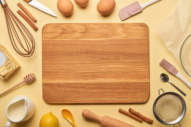 top view of empty wooden chopping board with cooking utensils and ingredients on yellow background  - Photo, Image
