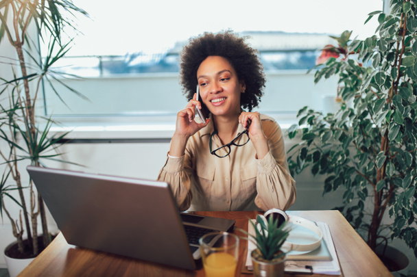 Sorridente giovane imprenditrice africana seduta a una scrivania nel suo ufficio di casa a lavorare online con un computer portatile
 - Foto, immagini