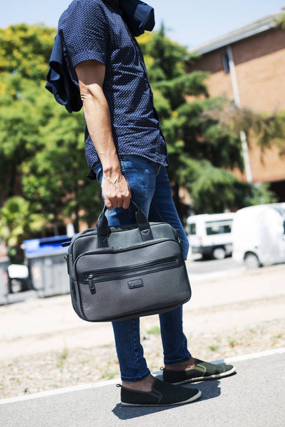 closeup of a young caucasian man, wearing a modern smart casual look, with slim jeans and untucked shirt, his jacket hanging in one shoulder, and carrying a briefcase, standing on the street - Fotoğraf, Görsel