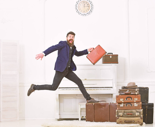 Macho attractive, elegant on cheerful face carries vintage suitcases, jumping. Man with beard and mustache in suit carries luggage, luxury white interior background. Long awaited vacation concept - Foto, Imagem