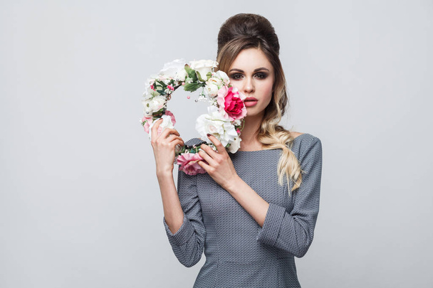 Portrait of beautiful attractive fashion model in grey dress with makeup and hairstyle holding flower wreath and looking at camera - Photo, Image