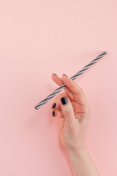 Creative top view flat lay of woman hand with black manicure holding cocktail drinking straw like cigarette with copy space pink paper background minimalism style. Template feminine blog social media - Фото, изображение