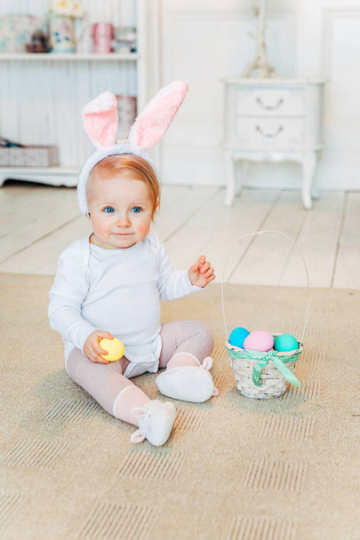 Pikkutyttö pupunkorvissa pääsiäispäivänä. Tyttö, jolla on kori, jossa on maalattuja munia, istuu lattialla kotona, hauskaa pääsiäismunan metsästyksessä. Hyvää pääsiäistä loma juhla kevät konsepti
 - Valokuva, kuva
