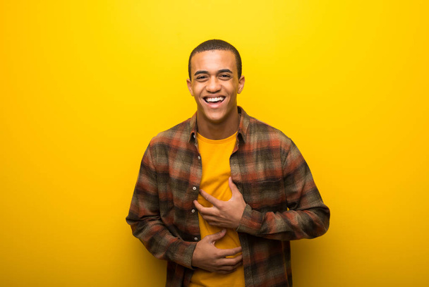 Молодий афроамериканець людиною на яскраві жовтому тлі багато посміхаючись при поклавши руки на грудях - Фото, зображення