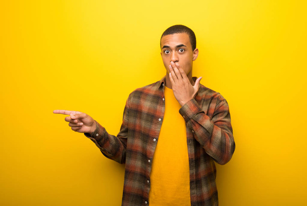 Fiatal afroamerikai férfi élénk sárga háttér mutató ujját, hogy az oldalán a meglepett arcot - Fotó, kép
