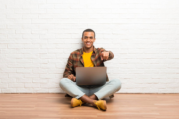 Afro-Américain assis sur le sol avec son ordinateur portable pointe du doigt vers vous avec une expression confiante
 - Photo, image