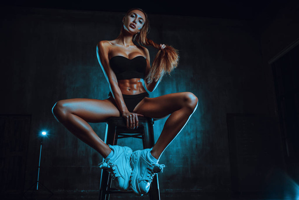 Young fitness woman sitting on chair in dark interior. Cool blue lights. - Foto, afbeelding