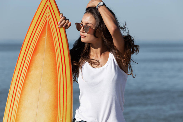 Beautiful slender dark-haired girl in sunglasses standing near by a  yellow surfboard  on the sandy beach near the sea. - Φωτογραφία, εικόνα