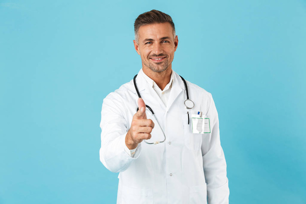 Confident man doctor wearing unifrom standing isolated over blue background, pointing at camera - Foto, Imagen