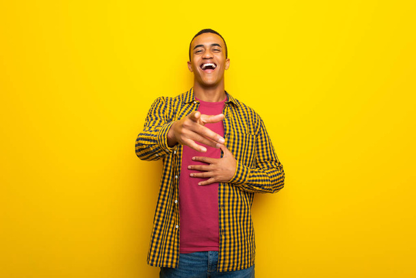 Nuori afroamerikkalainen mies keltaisella taustalla osoittaa sormella jotakuta ja nauraa paljon
 - Valokuva, kuva