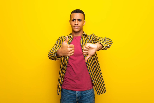 Afro Αμερικανός νεαρός σε κίτρινο φόντο κάνει καλό-κακό σημάδι. Αναποφάσιστοι μεταξύ Ναι ή όχι - Φωτογραφία, εικόνα