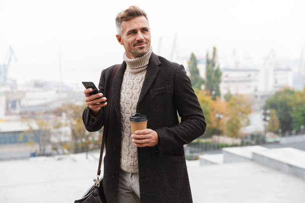 Portrait of attractive man 30s wearing jacket using mobile phone and holding takeaway coffee while walking through city street - Foto, imagen