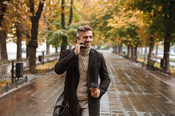 Photo of adult stylish man 30s wearing warm clothes walking outdoor through autumn park and using mobile phone - Zdjęcie, obraz