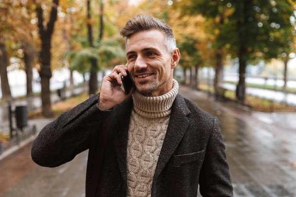 Photo of caucasian man 30s wearing warm clothes walking outdoor through autumn park and using mobile phone - Fotografie, Obrázek