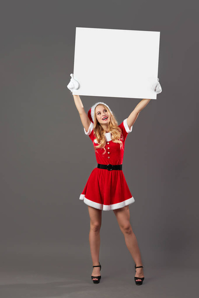 Young and beautiful mrs.Santa Claus dressed in the red robe, Santas hat, gloves  and high heels holds  the white canvas over her head on the gray background. - Photo, image