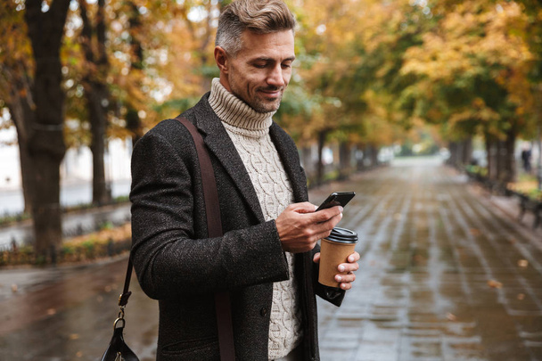 Photo of stylish macho man 30s wearing warm clothes walking outdoor through autumn park and using mobile phone - Photo, Image
