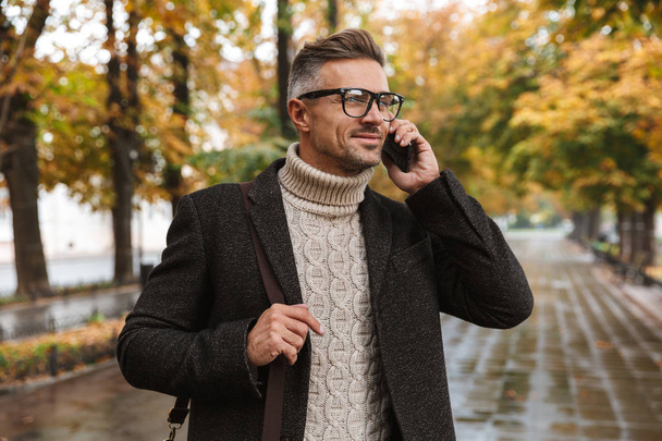 Photo of happy macho man 30s wearing warm clothes walking outdoor through autumn park and using mobile phone - Фото, изображение