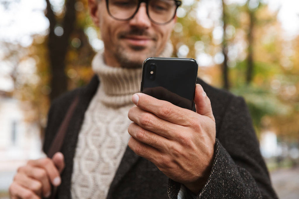 Foto de homem atraente 30 anos vestindo roupas quentes andando ao ar livre através do parque de outono e usando smartphone
 - Foto, Imagem